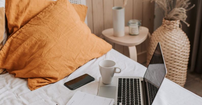 Affordable Home Decor - From above of open netbook with black screen and cup of coffee with smartphone and notebook placed on crumpled bed sheet in bedroom with big vase on floor