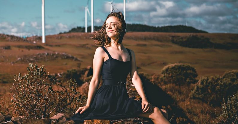 Industrial Chic - Side view of graceful young female ballet dancer in elegant dress and pointe shoes sitting on old stone fence with closed eyes near windmills in countryside